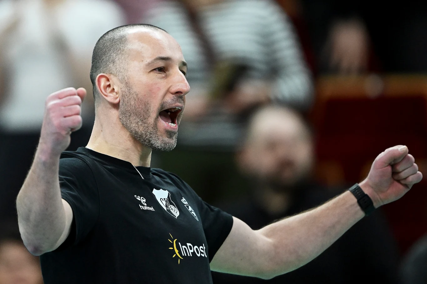 Krzysztof Andrzejewski, trener InPost ChKS Chełm Mężczyzna w czarnej koszulce z logo InPost, z uniesionymi w geście triumfu rękami, wyrażający silne emocje, prawdopodobnie podczas wydarzenia sportowego, na tle rozmytego tłumu.