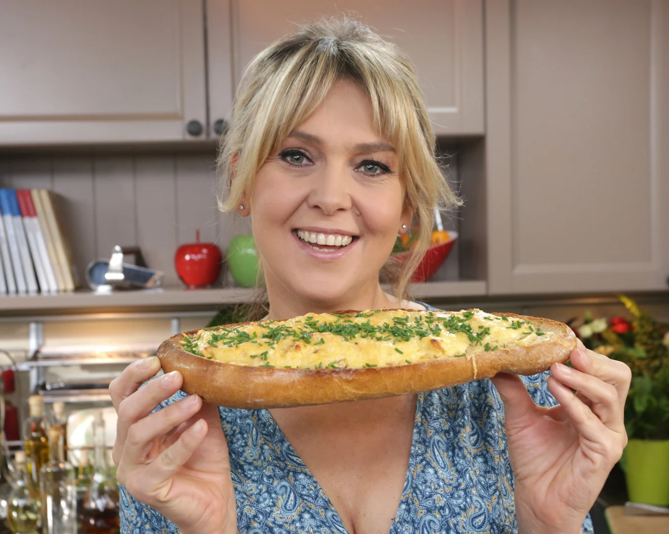 Uśmiechnięta kobieta w kuchni trzyma przed sobą dużą bagietkę zapiekaną z jajkami i posiekaną natką pietruszki, w tle widoczne są kuchenne półki oraz dekoracyjne elementy jak książki kucharskie i owoce.