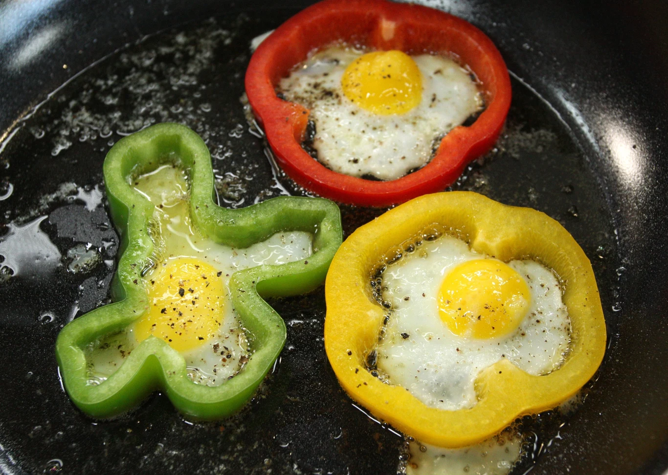 Kolorowe plasterki papryki w kształcie kwiatów i pierścieni, wypełnione smażonymi jajkami sadzonymi, leżące na rozgrzanej patelni z widocznym tłuszczem i przyprawami.