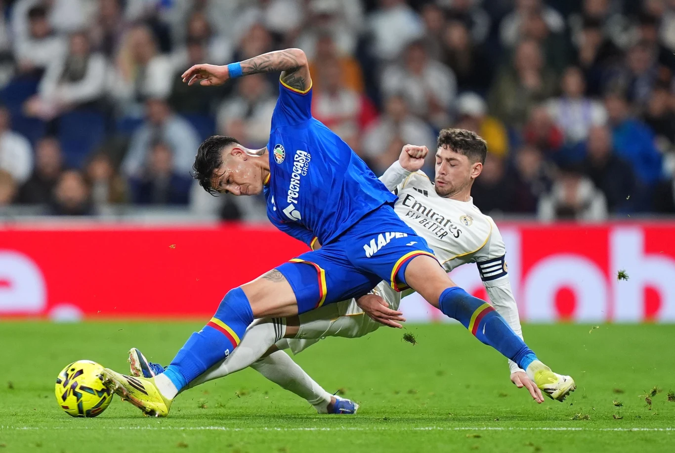 Dwóch piłkarzy walczy o piłkę na murawie podczas meczu, jeden gracz w niebieskim stroju wykonuje wślizg, drugi w białej koszulce i z opaską kapitańską próbuje przejąć piłkę, wokół rozpryskuje się trawa, w tle wyraźnie widoczni kibice na trybunach.