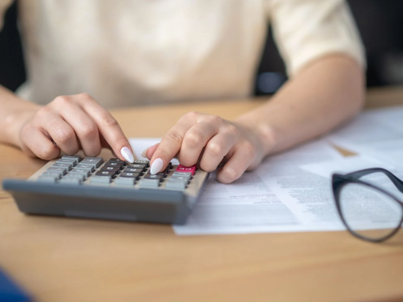 Dłonie osoby używającej kalkulatora na biurku z dokumentami oraz okularami leżącymi obok.