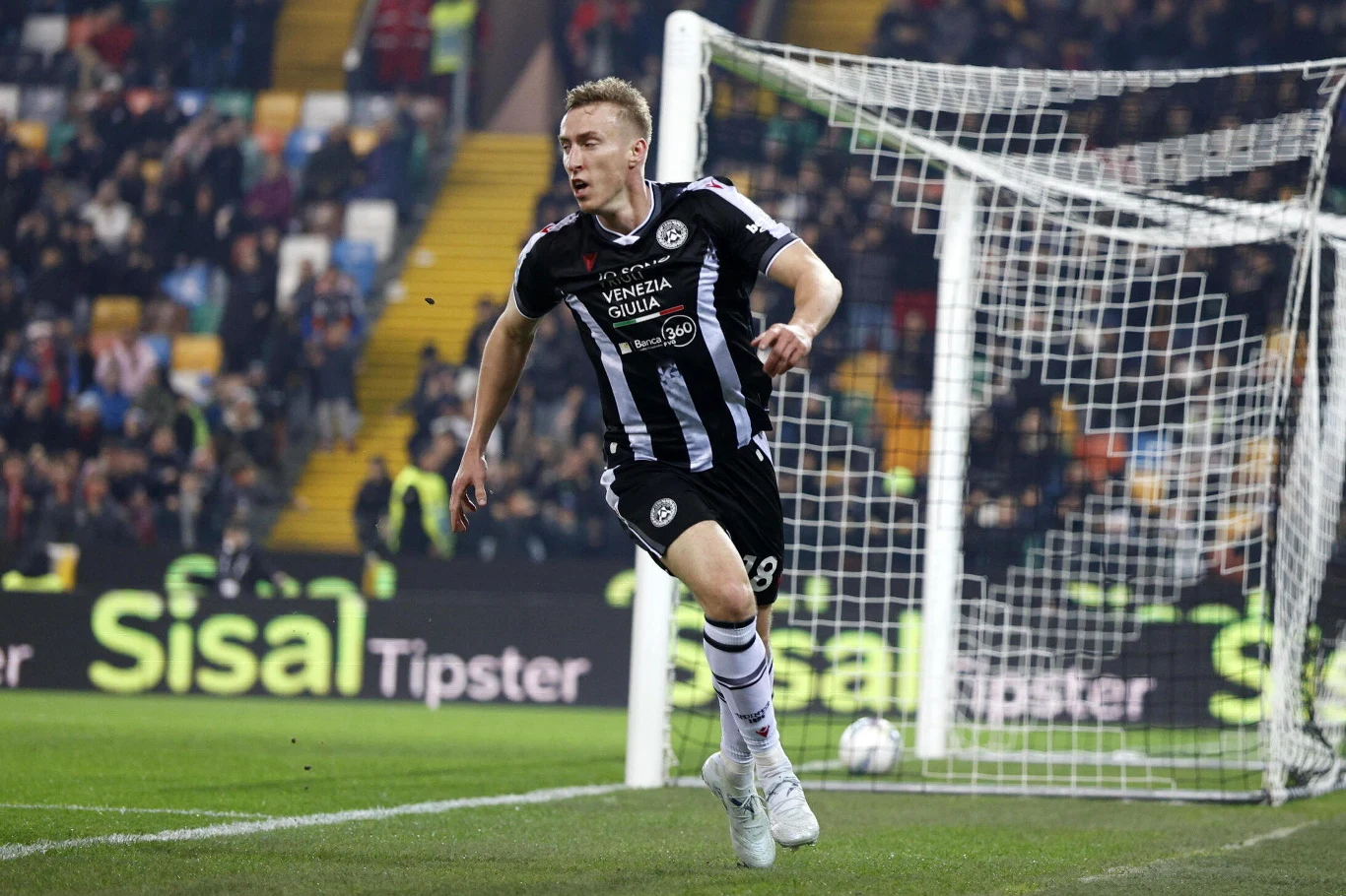Piłkarz w czarno-białym stroju drużyny Udinese biegnie dynamicznie po murawie w okolicy bramki podczas meczu piłkarskiego, na tle stadionu wypełnionego kibicami.