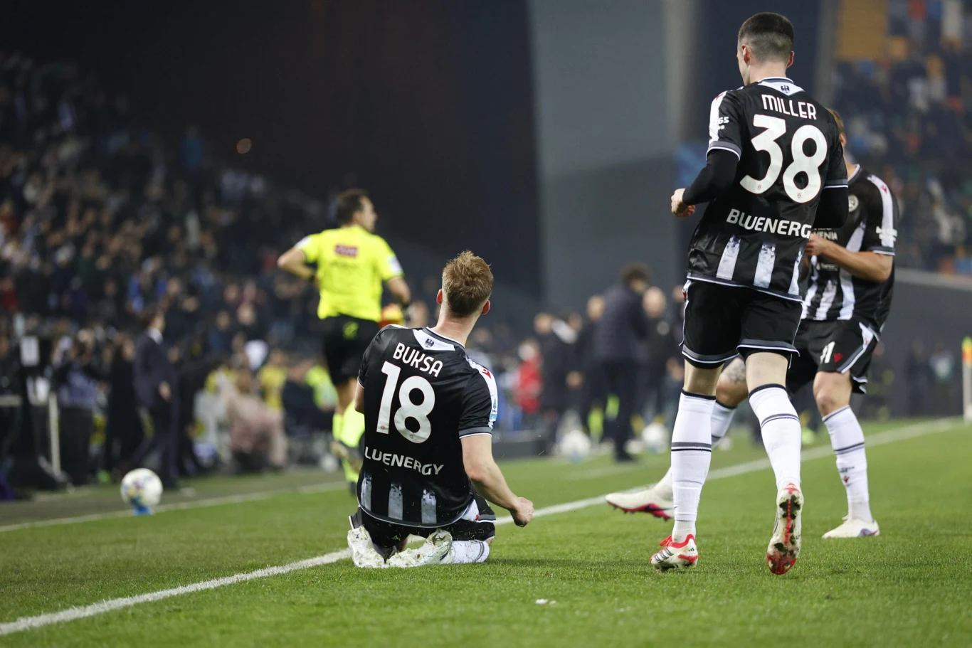 Piłkarz z numerem 18, siedzący na murawie obok linii bocznej, gestykuluje po akcji meczowej, podczas gdy inny zawodnik z numerem 38 podchodzi do niego; w tle widoczny sędzia biegnący oraz rozmyty tłum kibiców na trybunach.