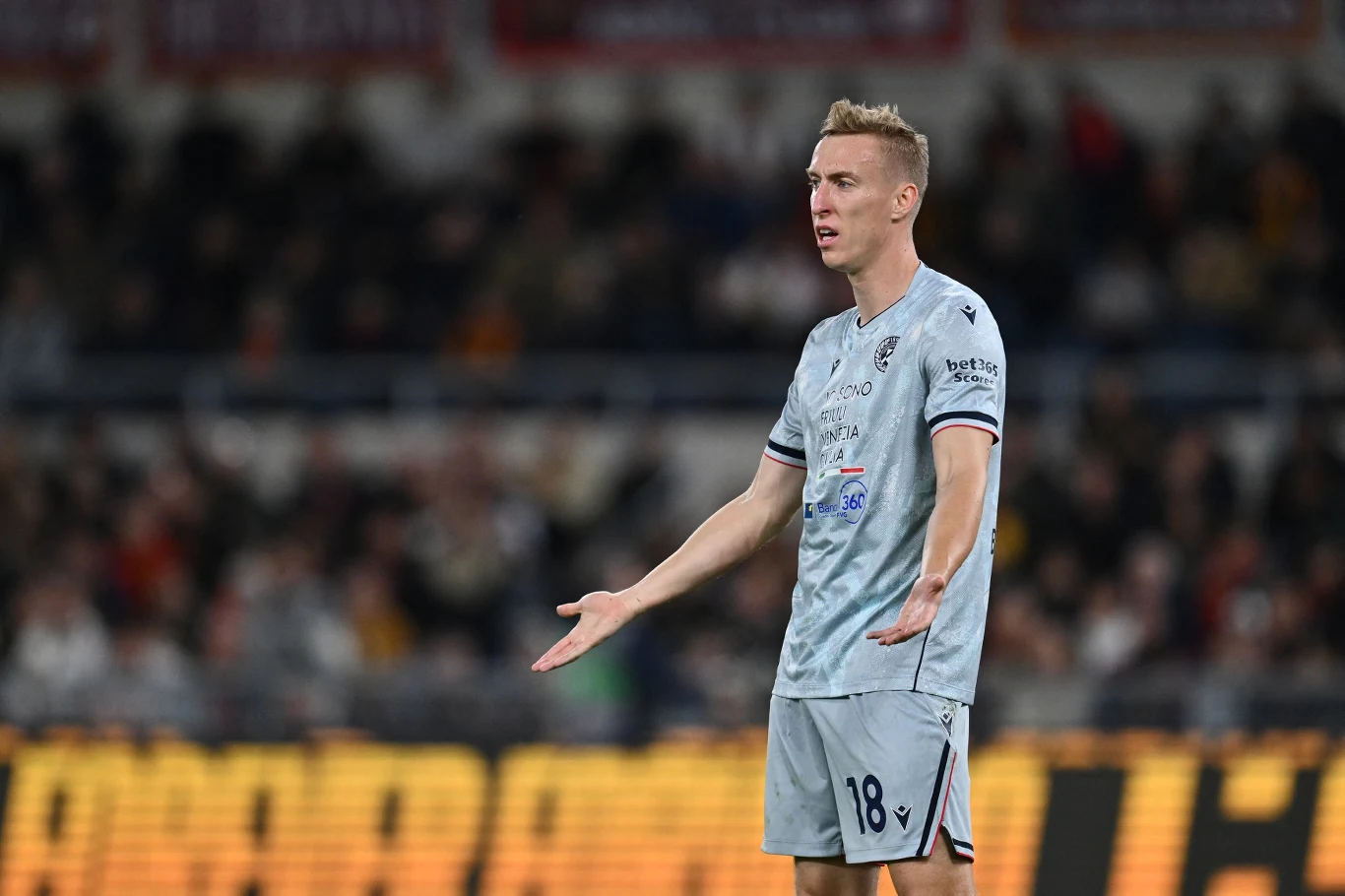 Piłkarz w jasnoszarym stroju z numerem 18 stoi na boisku z szeroko rozłożonymi rękami, wyrażając zaskoczenie lub dezaprobatę podczas meczu piłkarskiego; w tle widoczna rozmyta publiczność na trybunach.