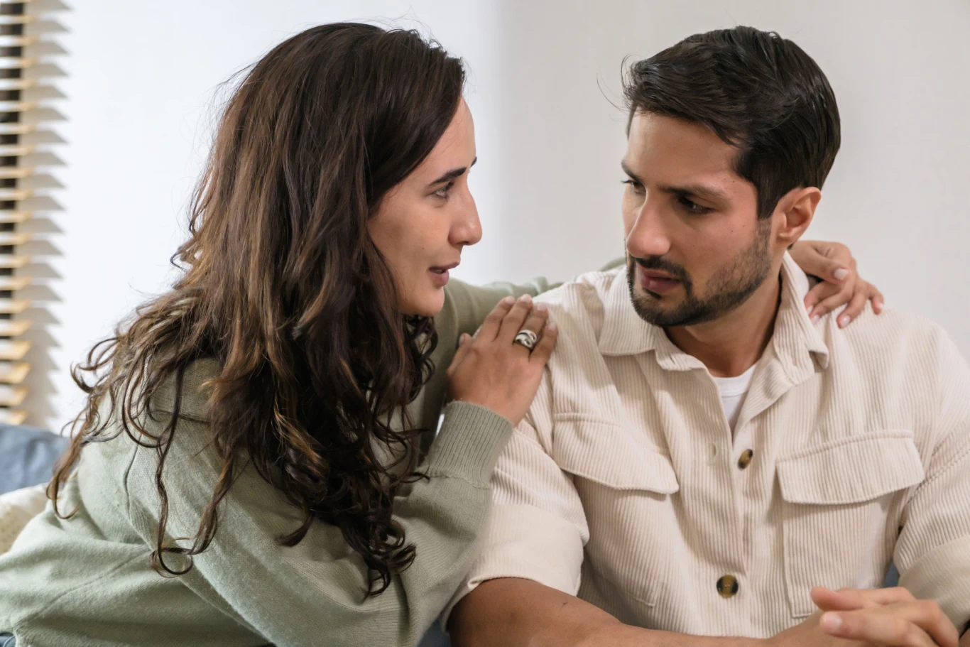 Kobieta z troską trzyma dłoń na ramieniu mężczyzny i patrzy mu w oczy, oboje prowadzą poważną rozmowę, wyrażając emocje zatroskania oraz wsparcia.