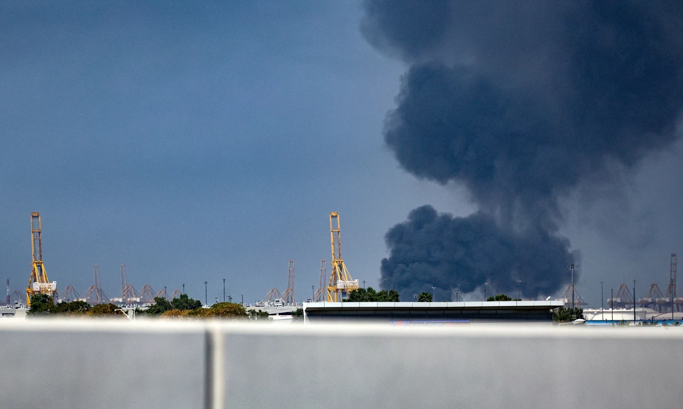 Odłamki pocisków spadły na port w Dżabal Ali w Dubaju, największym mieście Zjednoczonych Emiratów Arabskich Gęsty, ciemny dym unoszący się nad kompleksem przemysłowym z widocznymi dźwigami portowymi na pierwszym planie, sugerujący poważny pożar lub awarię.