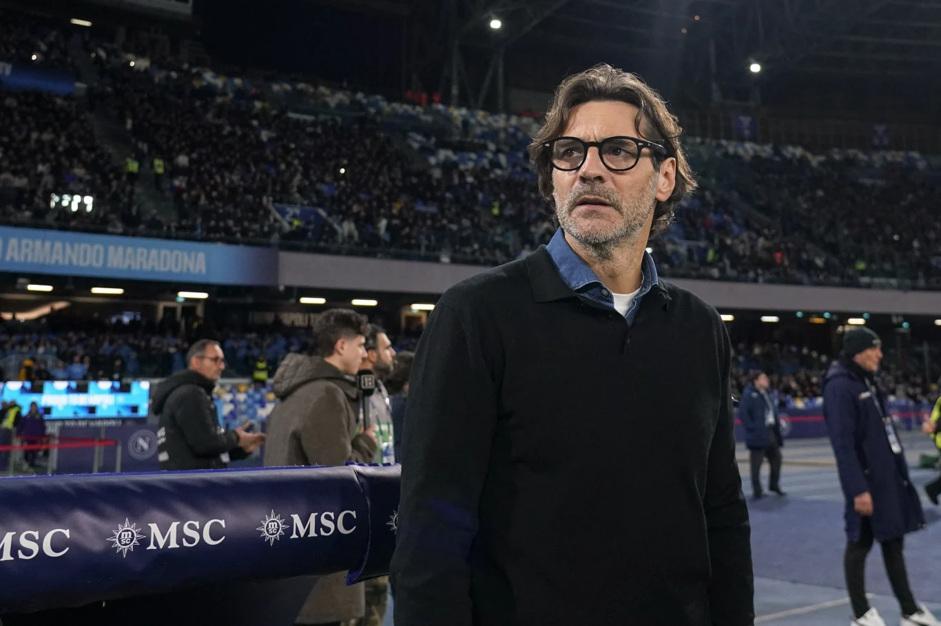 Paolo Vanoli Mężczyzna w okularach ubrany w czarny sweter stoi na stadionie piłkarskim, a w tle widoczny jest tłum kibiców oraz podświetlony baner ze słowem 'Maradona'.