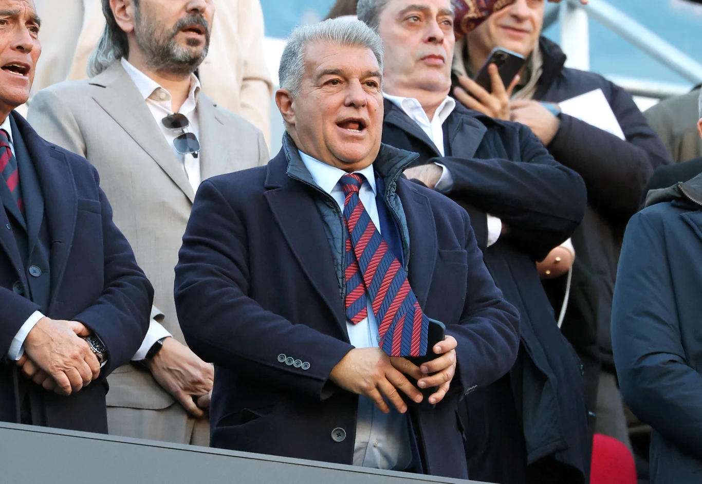 Elegancko ubrany mężczyzna w marynarce i krawacie w pasiasty wzór stoi wśród kilku innych osób, które także mają formalne stroje; w tle widoczne są fragmenty trybun stadionu.