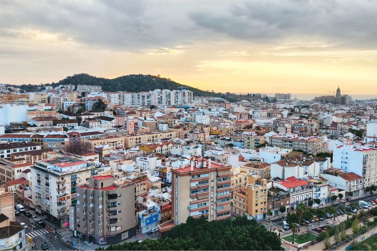 Panorama miasta z gęstą zabudową mieszkalną, licznymi budynkami o różnej wysokości, ulicami tworzącymi regularną siatkę i wzgórzem porośniętym zielenią w tle, przy świetle zachodzącego słońca.