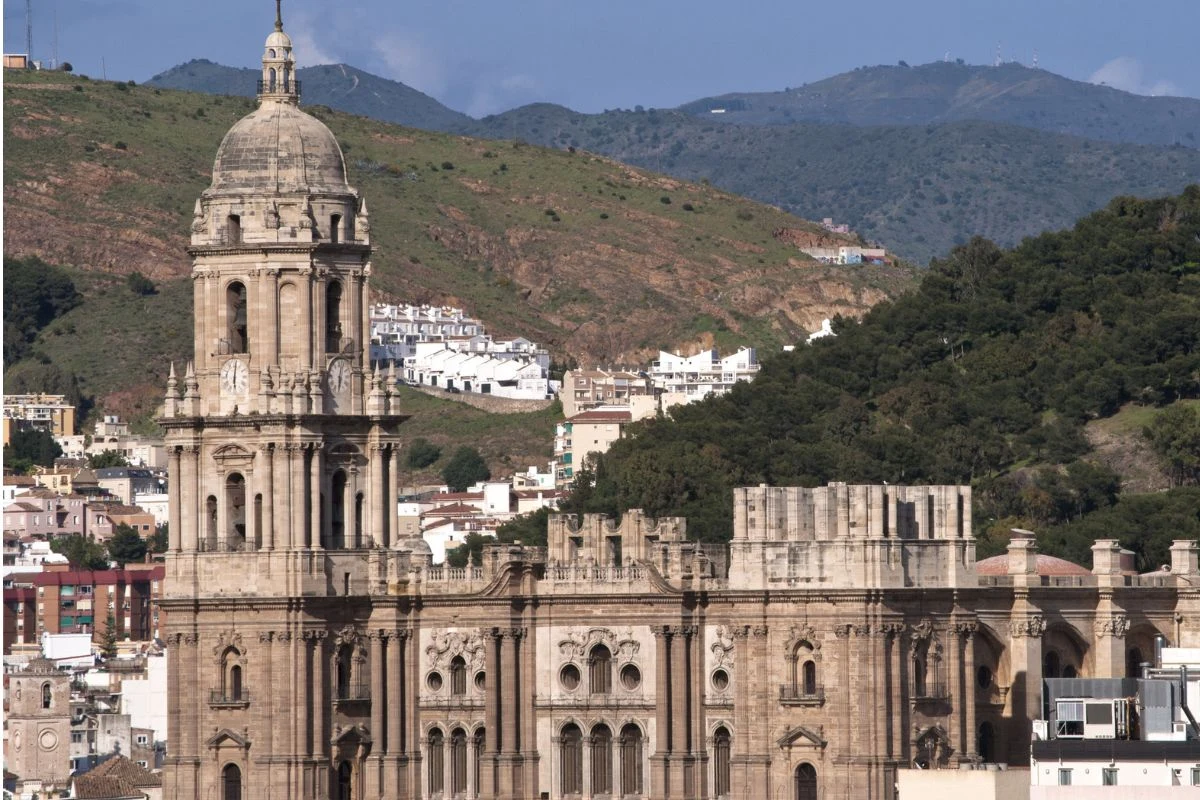 Katedra z wysoką kamienną wieżą na pierwszym planie, otoczona zielonymi wzgórzami oraz zabudowaniami mieszkalnymi w tle.