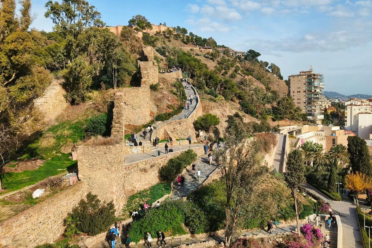 Zabytkowe mury miejskie pnące się po wzgórzu, liczni spacerowicze przemierzający ścieżkę wśród bujnej roślinności, w tle nowoczesne budynki miasta kontrastujące z historyczną architekturą.