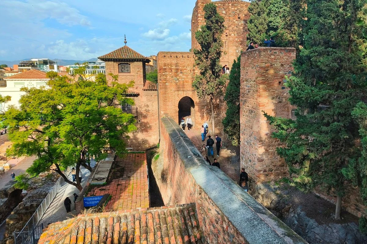 Stare kamienne mury fortecy, prowadząca przez nie ścieżka spacerowa otoczona drzewami oraz przechodzący ludzie, a w tle fragmenty historycznych zabudowań i błękitne niebo.