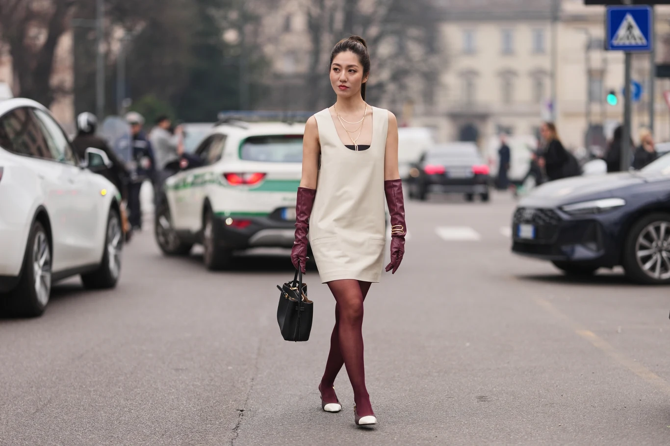 Kobieta idąca środkiem ulicy w białej sukience, bordowych rajstopach i długich bordowych rękawiczkach, z czarną torebką w dłoni; w tle widoczne samochody, w tym radiowóz policyjny, oraz kilka osób po obu stronach ulicy.