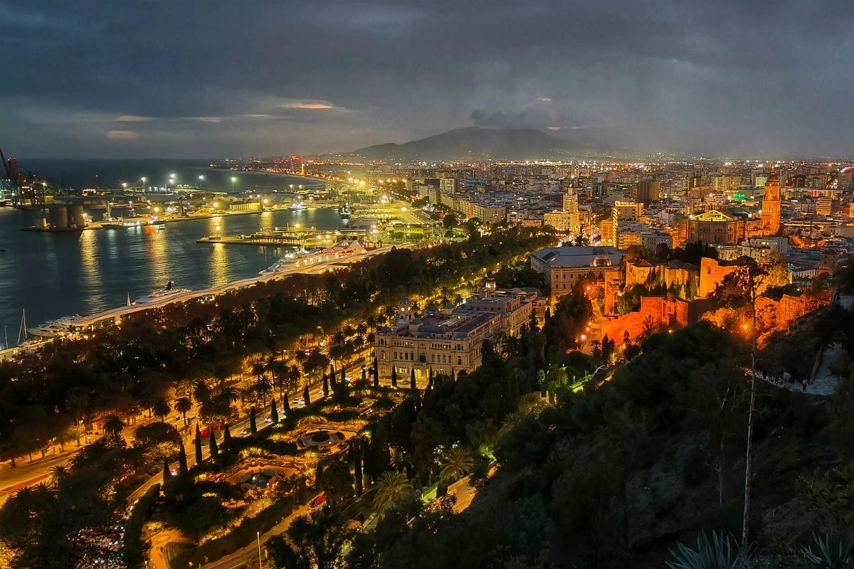 Panorama miasta o zmierzchu z oświetlonymi ulicami, portem nad morzem po lewej stronie oraz zabytkowymi budynkami z wyraźnie podświetlonym zamkiem i katedrą na pierwszym planie.