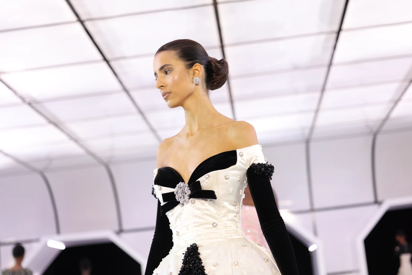 Kobieta w eleganckiej sukni wieczorowej z czarno-białymi elementami oraz rękawiczkami, uczesana w gładki kok, prezentuje się na futurystycznym wybiegu o jasnym, geometrycznym suficie.