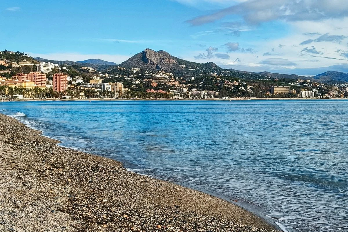 Szeroka, kamienista plaża nad spokojnym morzem, w tle panorama miasta z zabudowaniami i wzgórzami, górującymi nad okolicą.