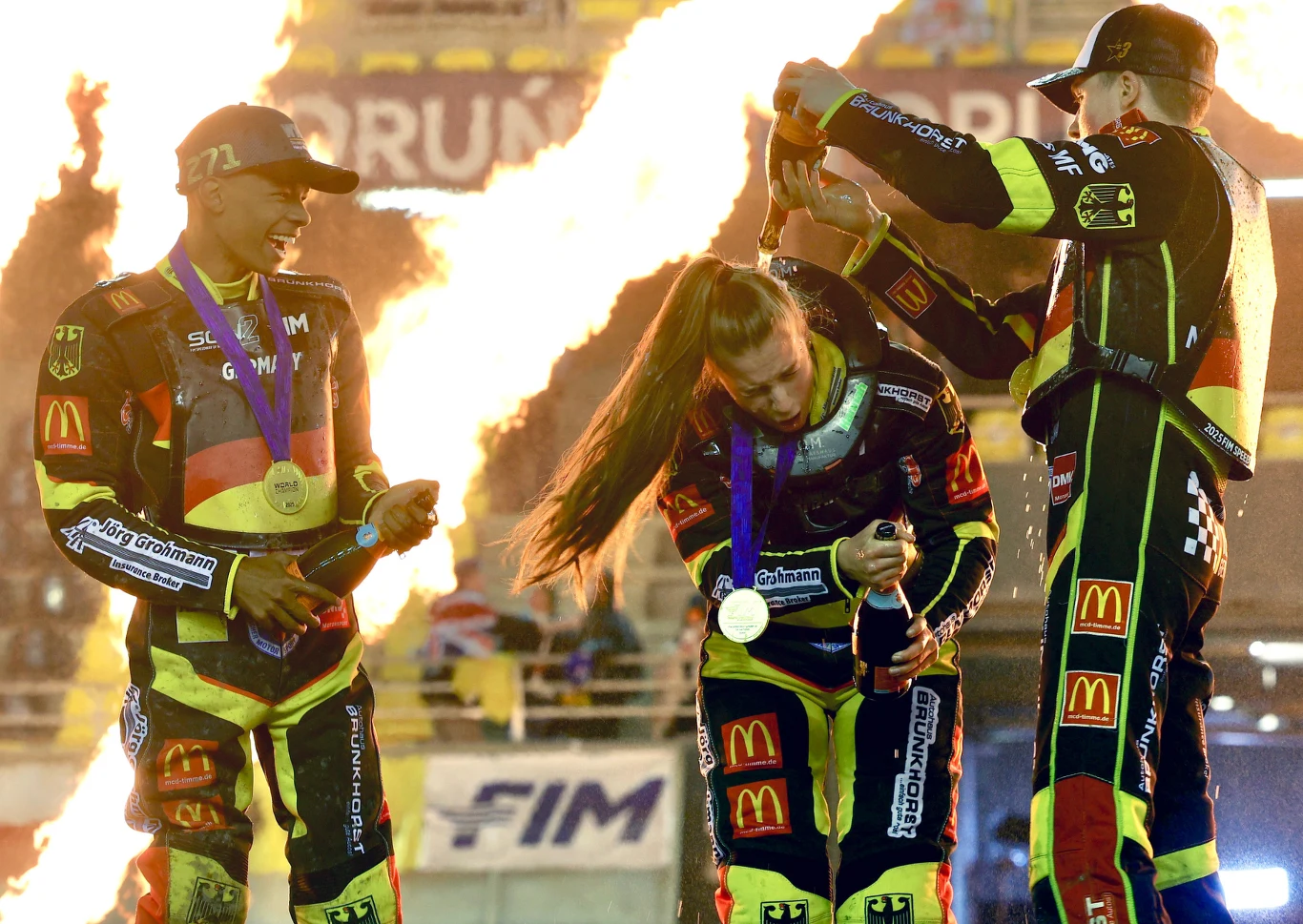 Troje sportowców w kombinezonach żużlowych z medalami na szyi świętuje zwycięstwo na stadionie, w tle widać wybuchy ognia i trybuny pełne światła.