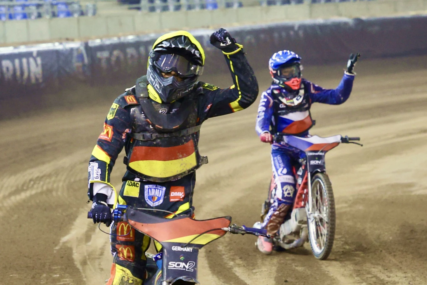 Dwóch żużlowców w kolorowych kombinezonach jedzie po torze, jeden z nich unosi zaciśniętą pięść w geście triumfu, drugi macha ręką, wokół nich widoczne trybuny stadionu.