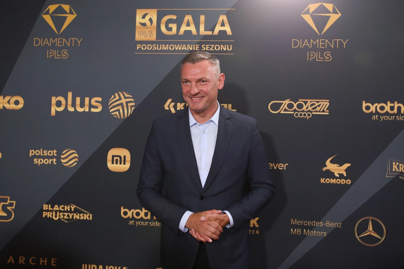 Mężczyzna w eleganckim garniturze stoi na tle ścianki z logotypami partnerów i sponsorów wydarzenia, związanej z galą podsumowania sezonu sportowego.