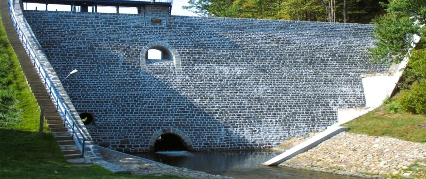 Wielkie zbrojenie w Jarnołtówku. Tak zmieni się zapora, która chroniła podczas powodzi w 2024 roku Kamienna zapora wodna w Jarnołtówku z okrągłymi otworami, otoczona zielenią i schodami na konstrukcji