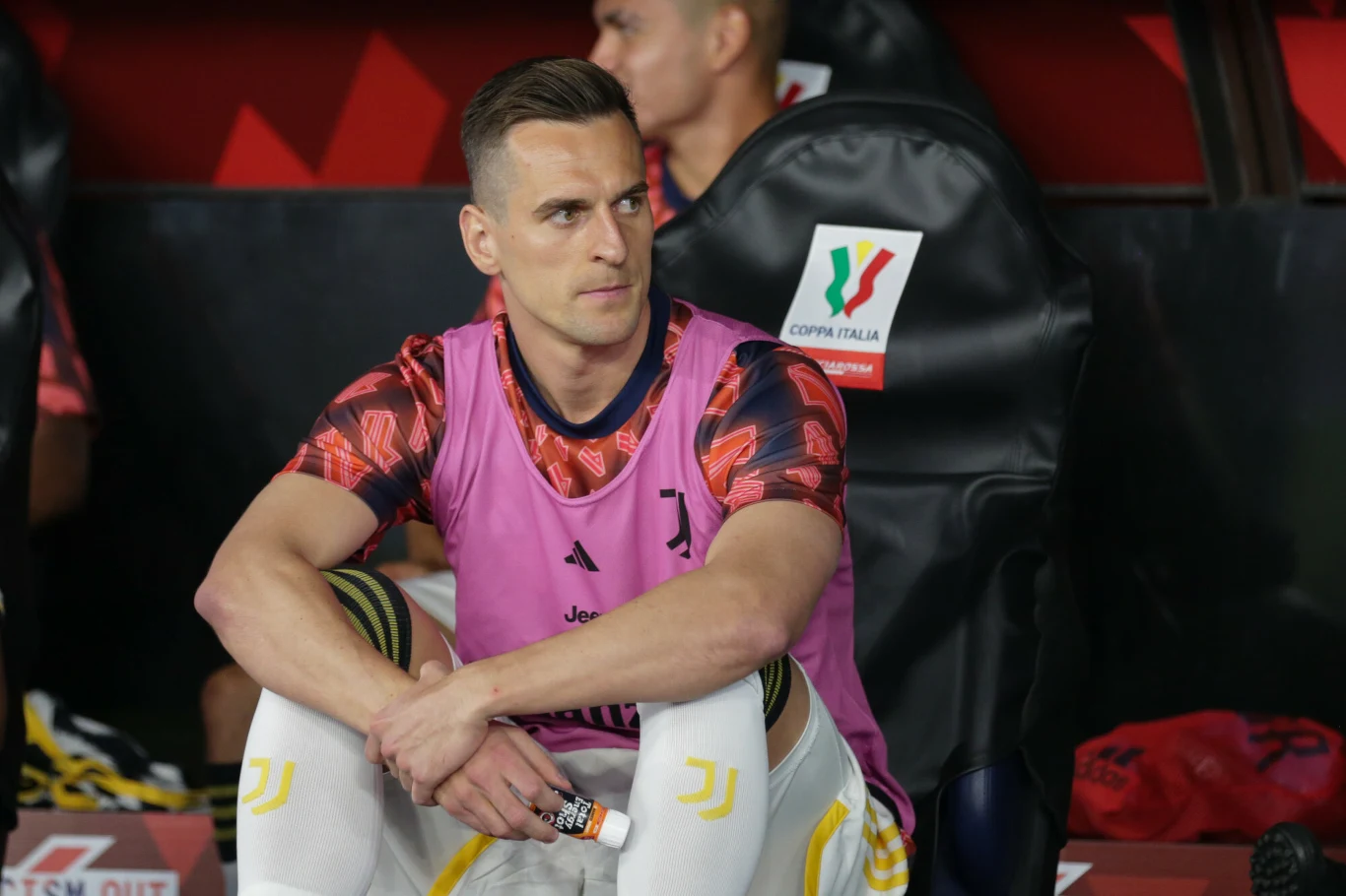 Piłkarz w stroju treningowym Juventusu siedzi na ławce rezerwowych, ubrany w różową kamizelkę i trzymający przedmiot w rękach, z widocznym emblematem Coppa Italia na oparciu krzesła za nim.