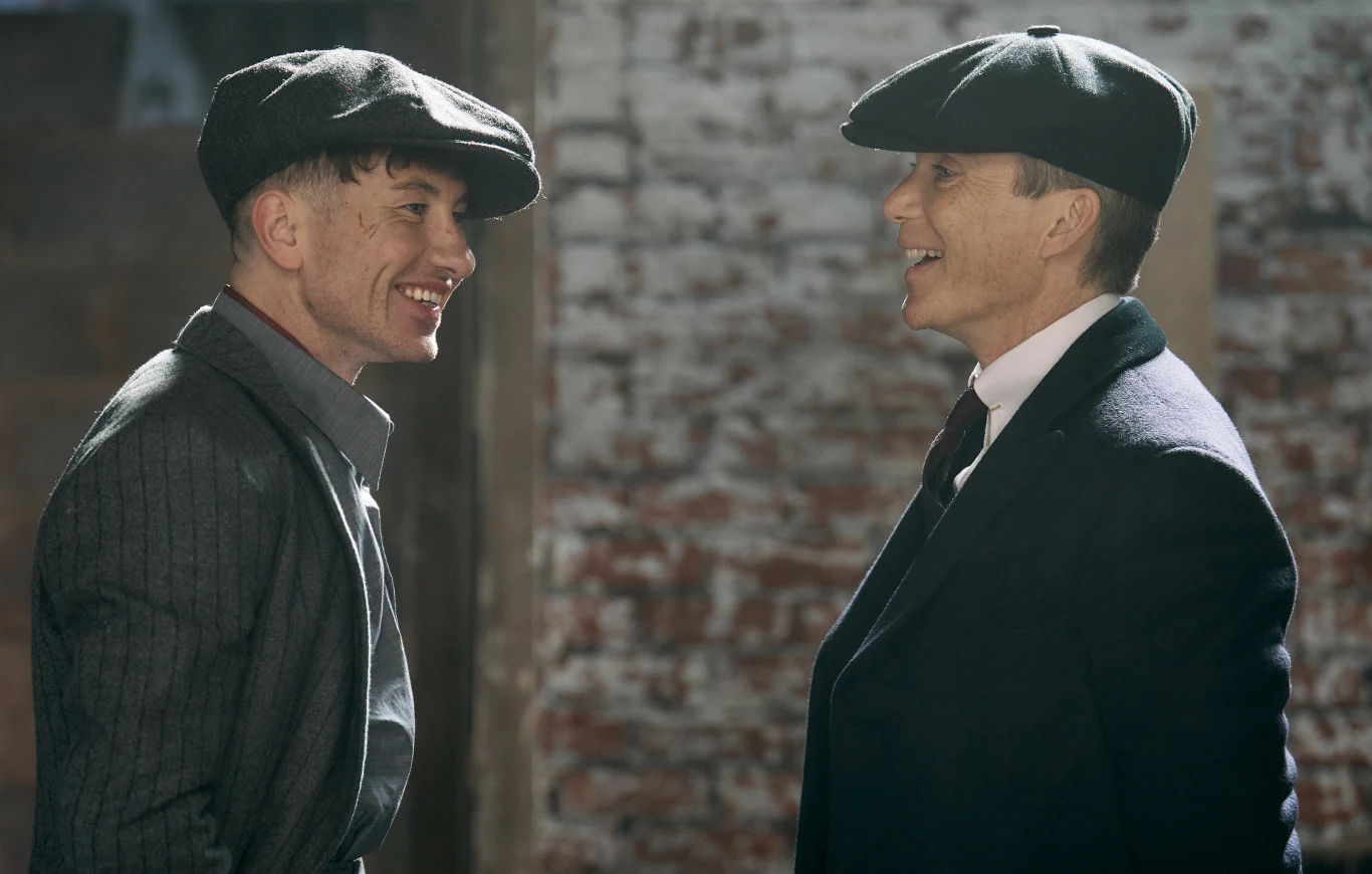 Dwóch uśmiechniętych mężczyzn w klasycznych garniturach i kaszkietach stoi twarzą w twarz na tle ceglanej ściany, prowadząc rozmowę.