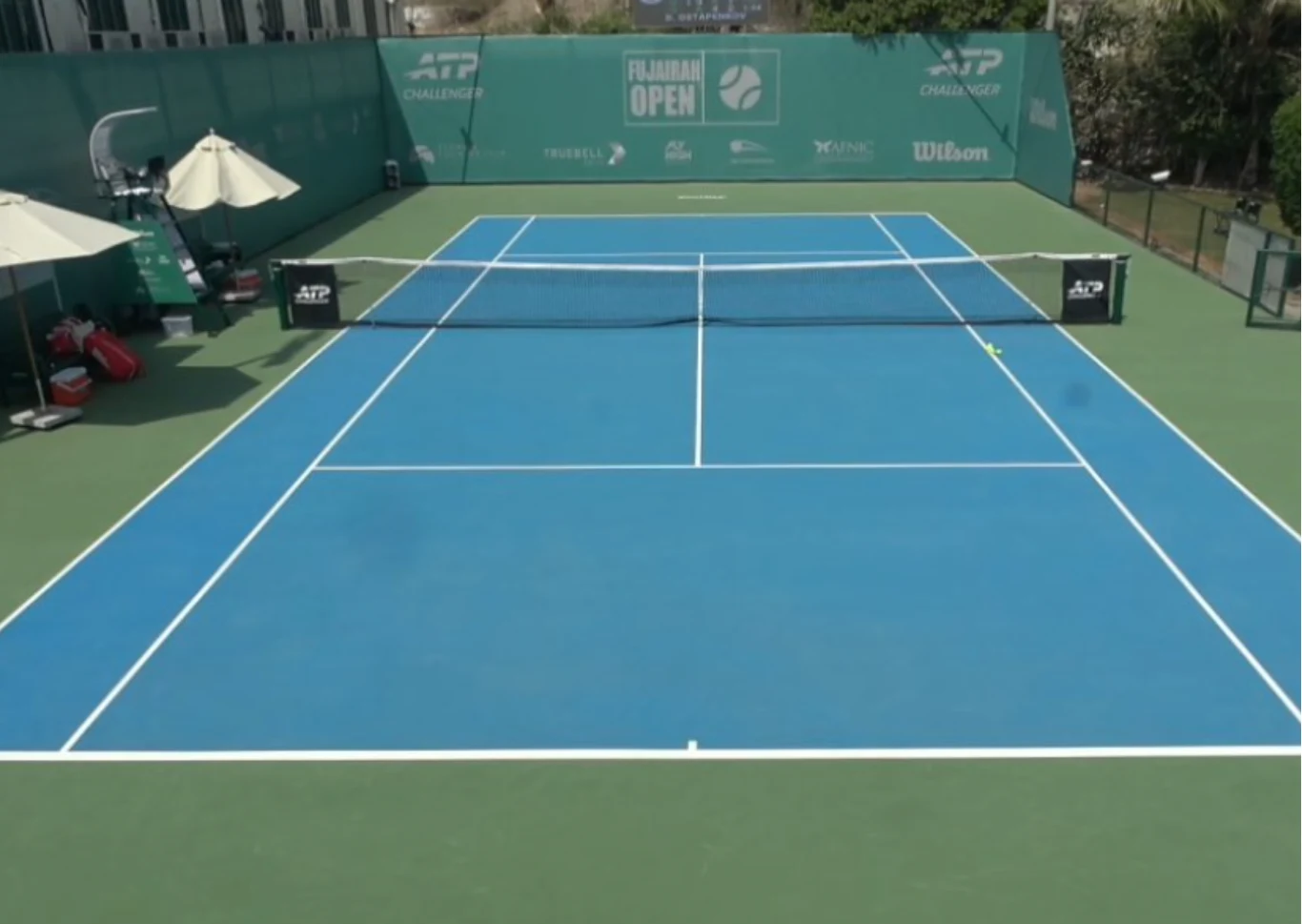 Pusty kort tenisowy o niebieskiej nawierzchni z siatką na środku, zielonymi bandami reklamowymi wokół i sprzętem sportowym ustawionym po lewej stronie wraz z parasolami.
