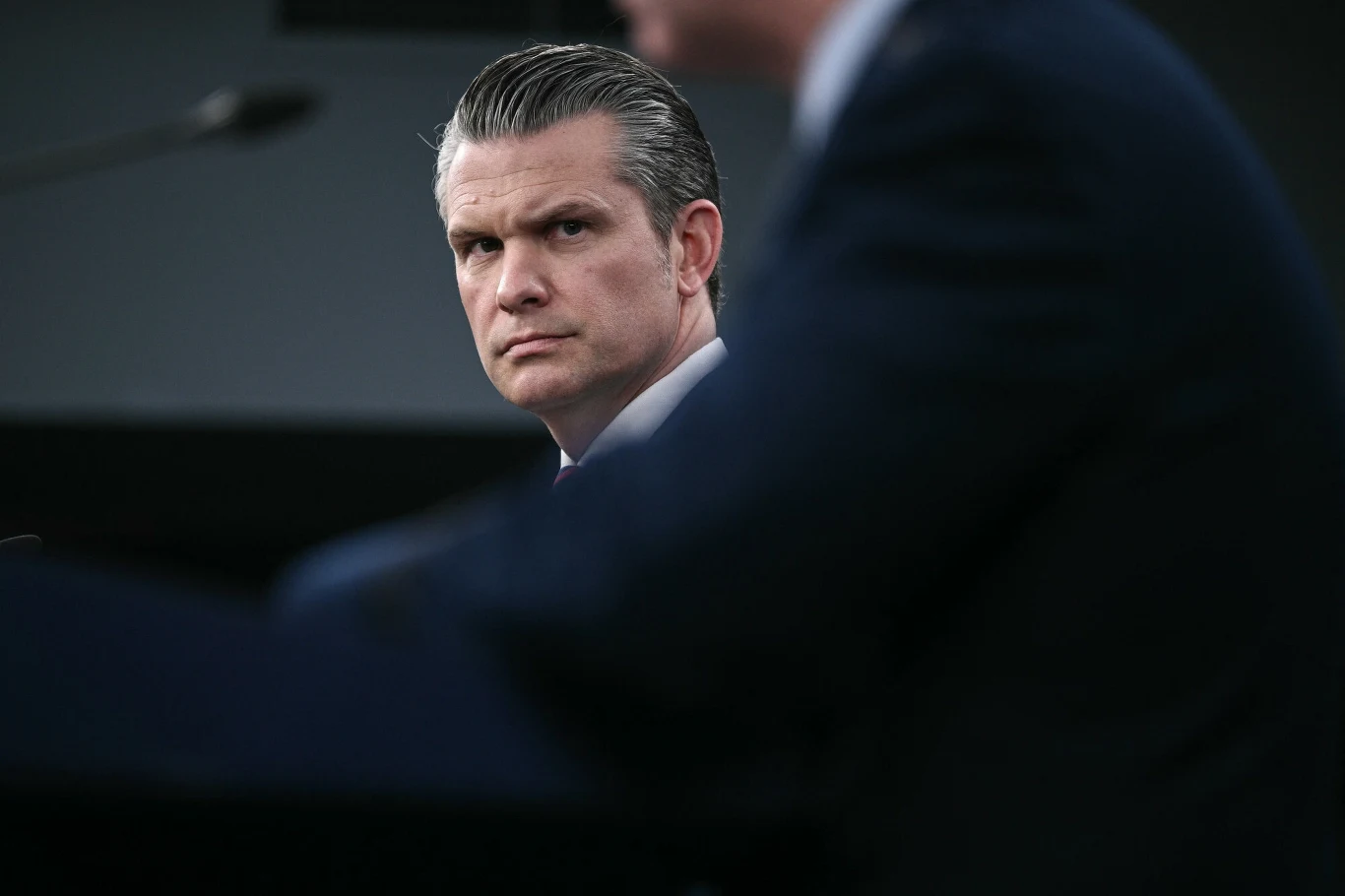 Mężczyzna w garniturze z poważnym wyrazem twarzy siedzi przy stole konferencyjnym, skupiając wzrok na rozmówcy znajdującym się w pierwszym planie.