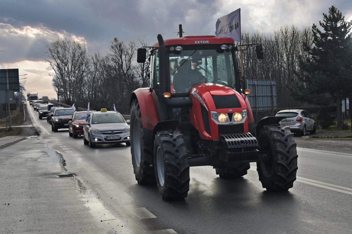 Czerwony traktor jadący po asfalcie prowadzi kolumnę samochodów osobowych, które poruszają się w jednym kierunku wśród zimowego krajobrazu z gołymi drzewami i zachmurzonym niebem.