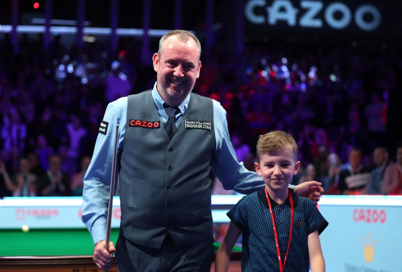 Mark Wiliams z synem podczas jednego z turniejów. 2023 rok Mężczyzna w eleganckim kamizelce i koszuli trzyma kij bilardowy, obok niego stoi uśmiechnięty chłopiec z zawieszoną na szyi czerwoną smyczą; oboje znajdują się na arenie snookerowej otoczonej przez widownię i oświetlonej kolorowymi światłami.