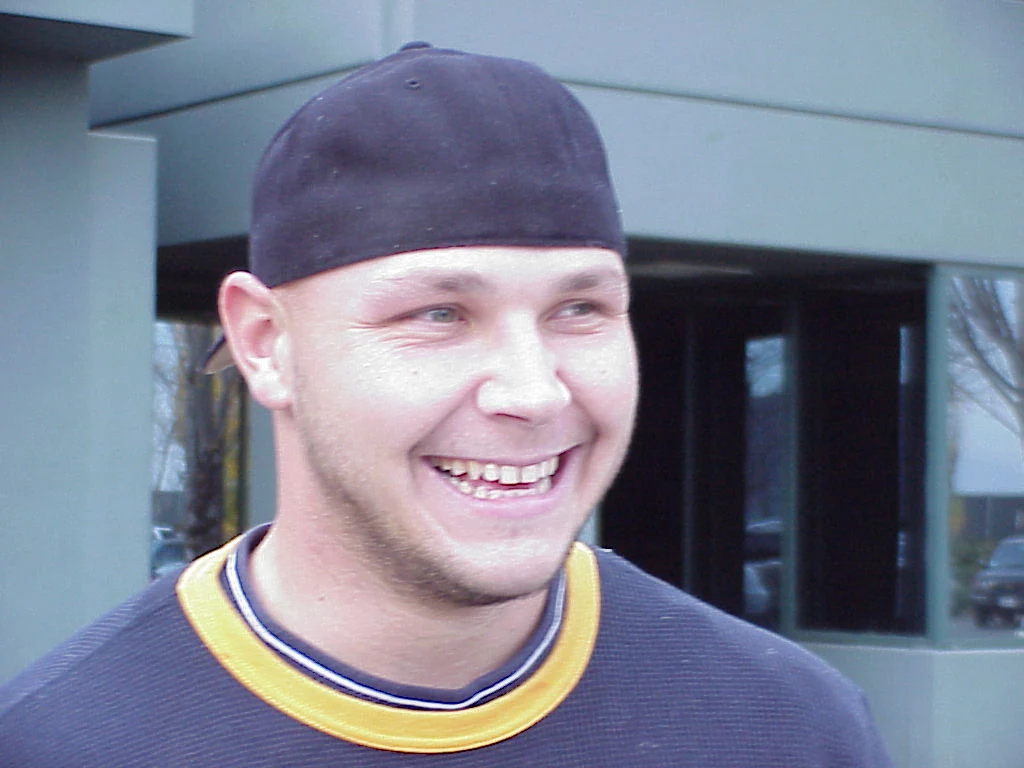 Sebastian Janikowski Młody mężczyzna w ciemnej czapce z daszkiem założonej tył do przodu i granatowej bluzie z żółtym kołnierzem uśmiecha się szeroko, stojąc przed szarym nowoczesnym budynkiem.