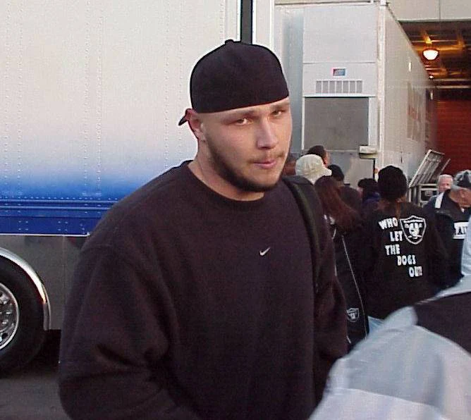 Sebastian Janikowski Mężczyzna w czarnej bluzie i czapce typu bandana stoi na tle białej ciężarówki, za nim grupa ludzi, z których jedna osoba ma na plecach napis 'Who let the dogs out'.