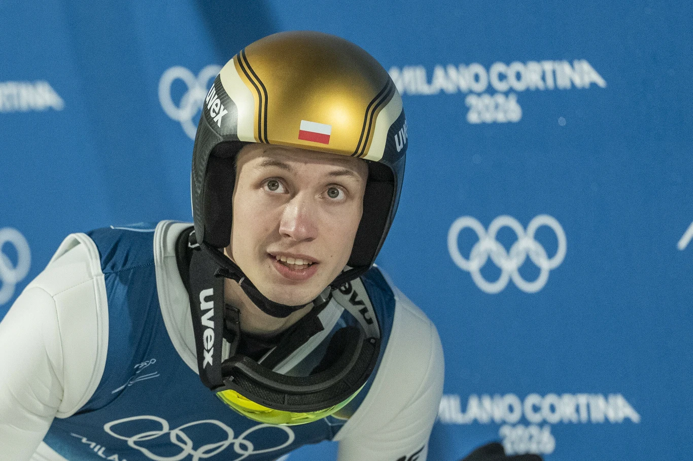 Sportowiec w kombinezonie startowym i złotym kasku z polską flagą, na tle niebieskiej ścianki z logo igrzysk olimpijskich Milano Cortina 2026.