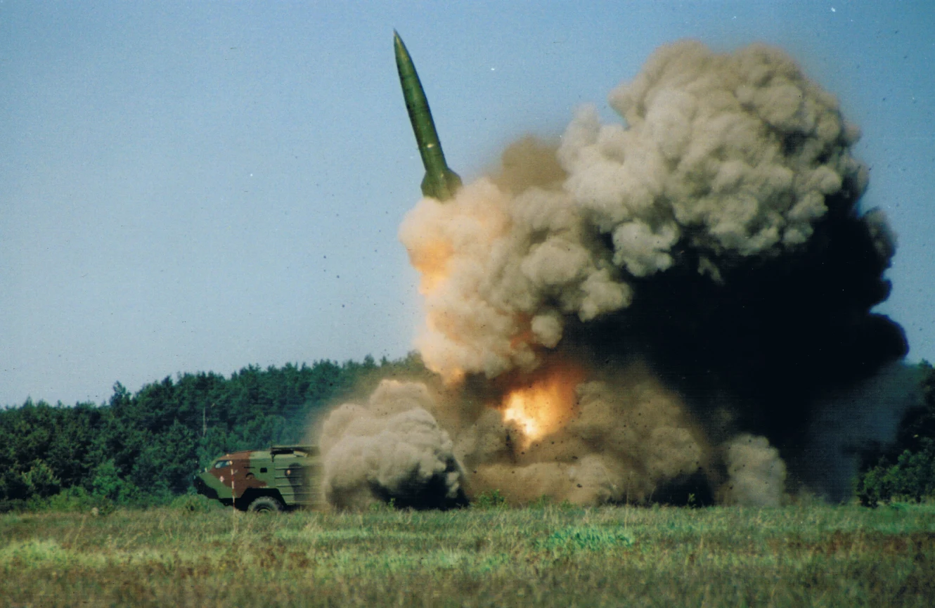 Pocisk balistyczny startujący z mobilnej wyrzutni, otoczony dużą chmurą dymu i pyłu na tle zieleni i lasu.