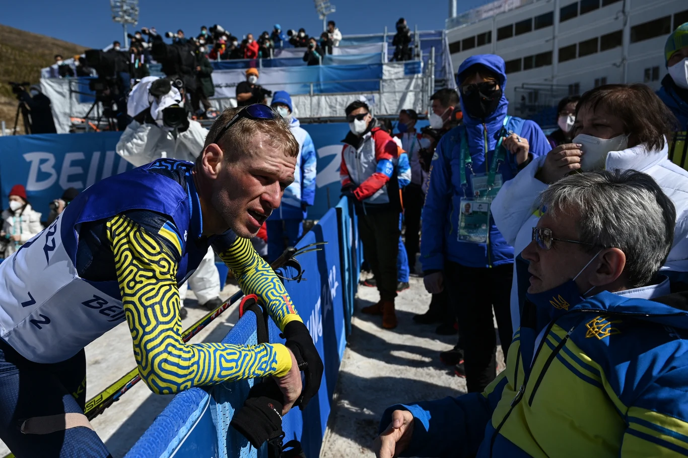 Zawodnik w sportowym stroju zimowym oparty o barierkę rozmawia z siedzącym mężczyzną w kurtce z emblematem, wokół nich znajduje się grupa osób w maseczkach obserwujących sytuację, w tle widoczne zaplecze sportowe i trybuny.