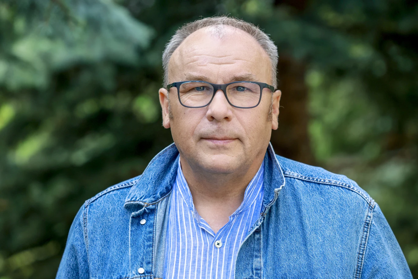 Mężczyzna w okularach ubrany w niebieską kurtkę jeansową i koszulę w jasnoniebieskie paski, patrzy prosto przed siebie na tle zielonych drzew.