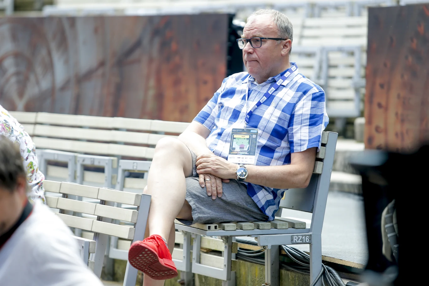 Mężczyzna w okularach, ubrany w niebiesko-białą koszulę w kratę, szare szorty i czerwone buty, siedzi na ławce z założoną nogą na nogę w pustym amfiteatrze.