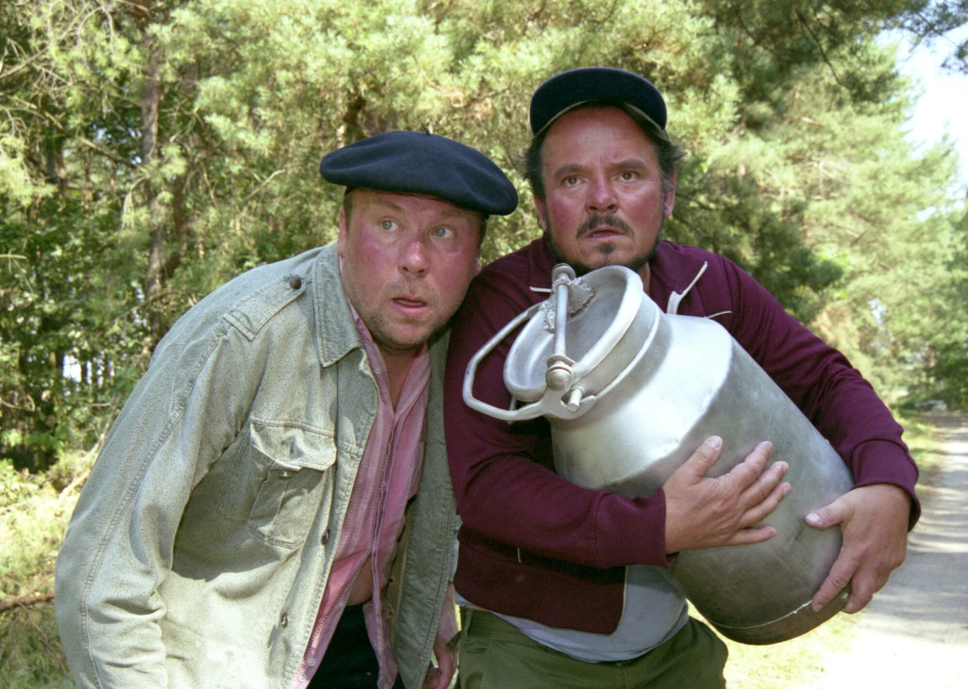 Dwóch mężczyzn w kaszkietach, jeden ubrany w jasną kurtkę, drugi w bordową bluzę, trzymających dużą metalową bańkę na mleko; obaj patrzą z niepokojem w jednym kierunku, stojąc na leśnej drodze.