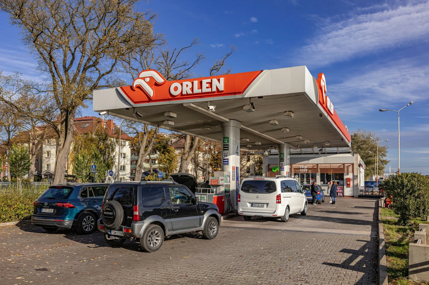 Stacja paliw Orlen z czterema samochodami zaparkowanymi przy dystrybutorach, otwartą klapą bagażnika w jednym z aut oraz widoczną ladą sklepu po prawej stronie pod zadaszeniem.