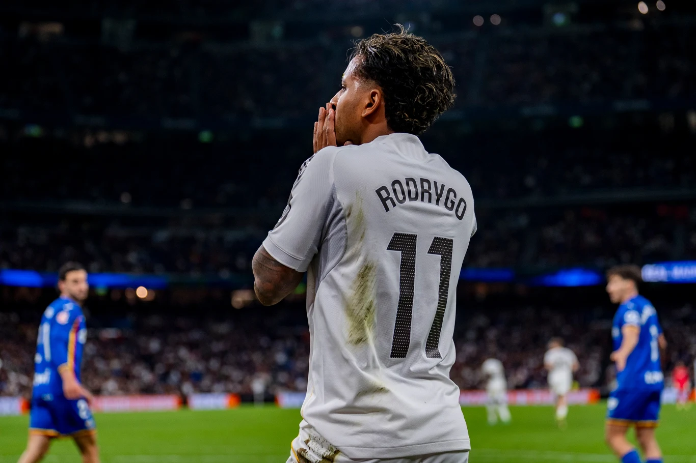 Rodrygo w białej koszulce z numerem 11, składa ręce przy twarzy, na tle stadionu z kibicami i innymi piłkarzami.