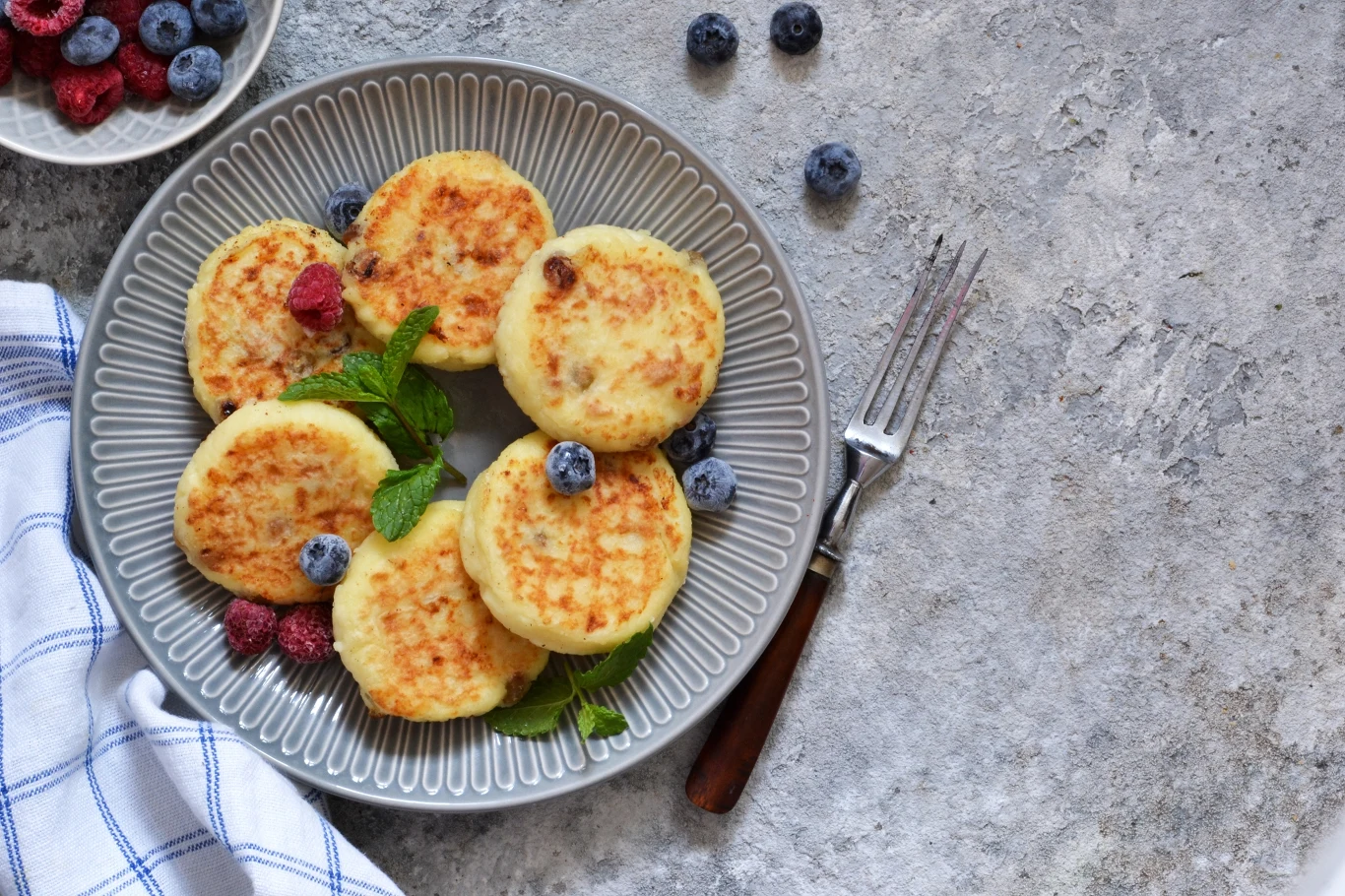 Sześć placków twarogowych ułożonych na szarym talerzu, ozdobionych świeżymi malinami, borówkami oraz listkami mięty, obok widelec i biała ściereczka w niebieską kratkę.