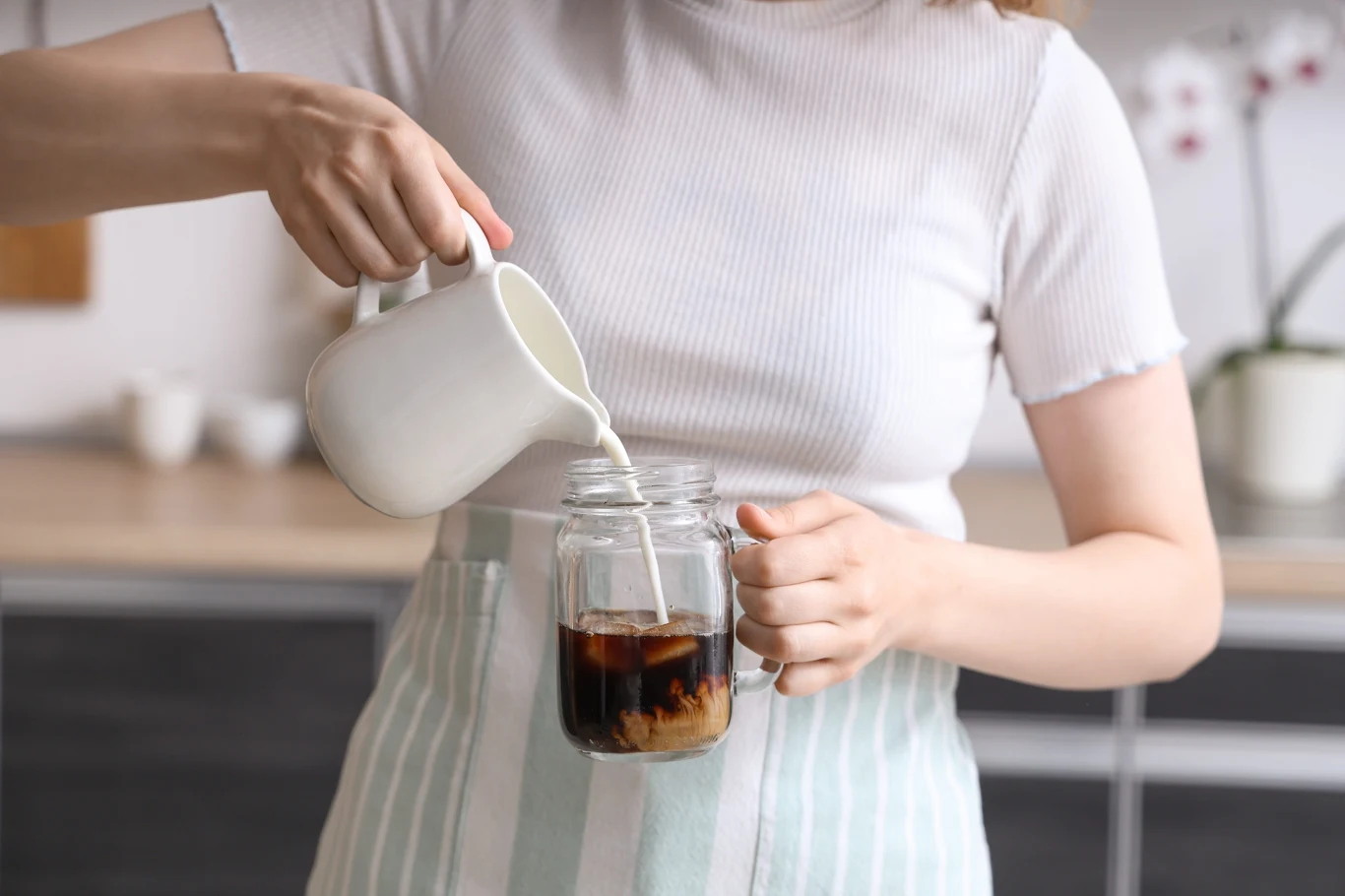 Młoda kobieta w jasnej bluzce i fartuchu wlewa mleko z białego dzbanka do dużej szklanki z kawą na tle nowoczesnej kuchni.