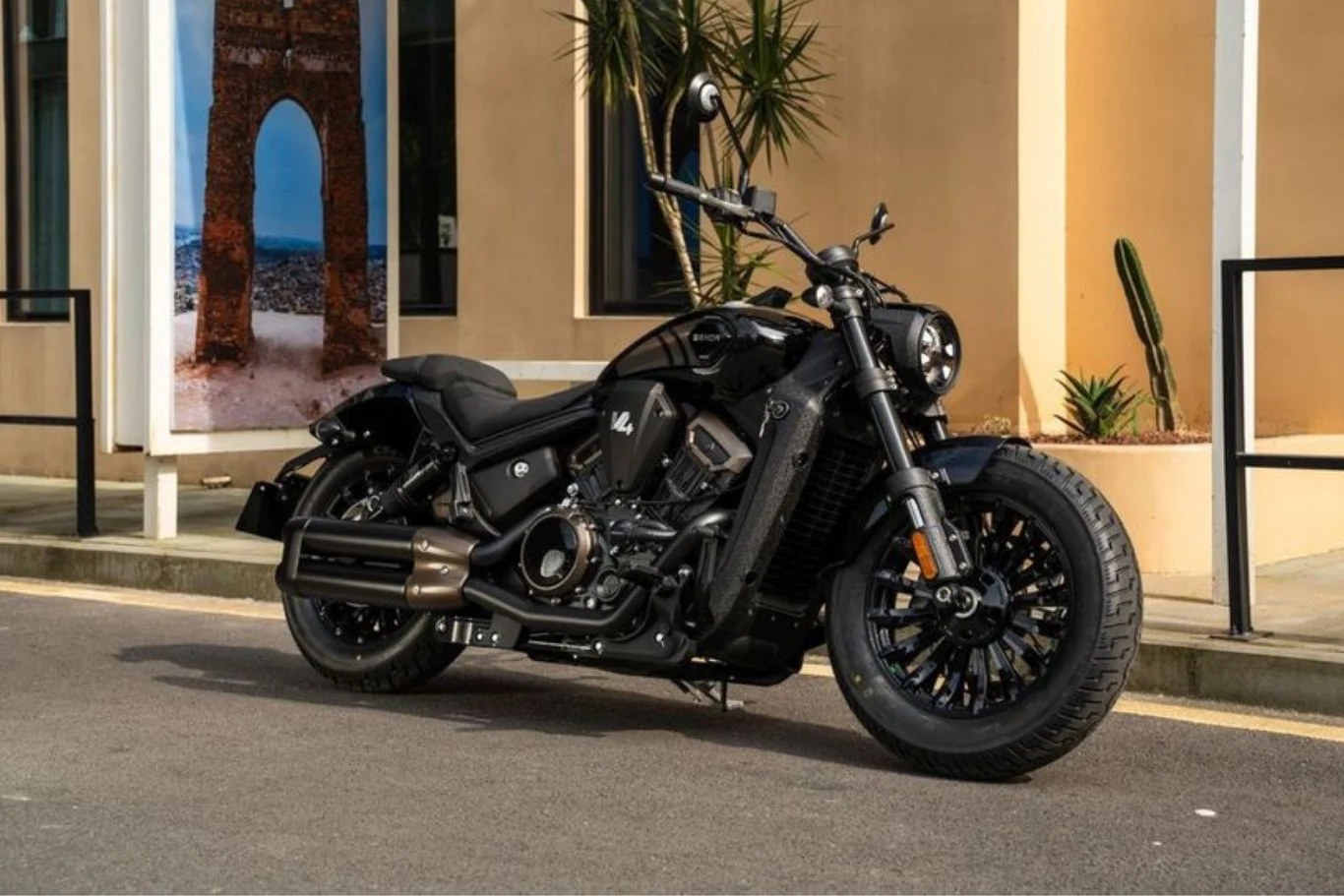 Nowoczesny, czarny motocykl o masywnej sylwetce zaparkowany na ulicy, z szerokimi oponami i matowym wykończeniem, w tle kremowy budynek z dużym oknem oraz wysokimi roślinami doniczkowymi.