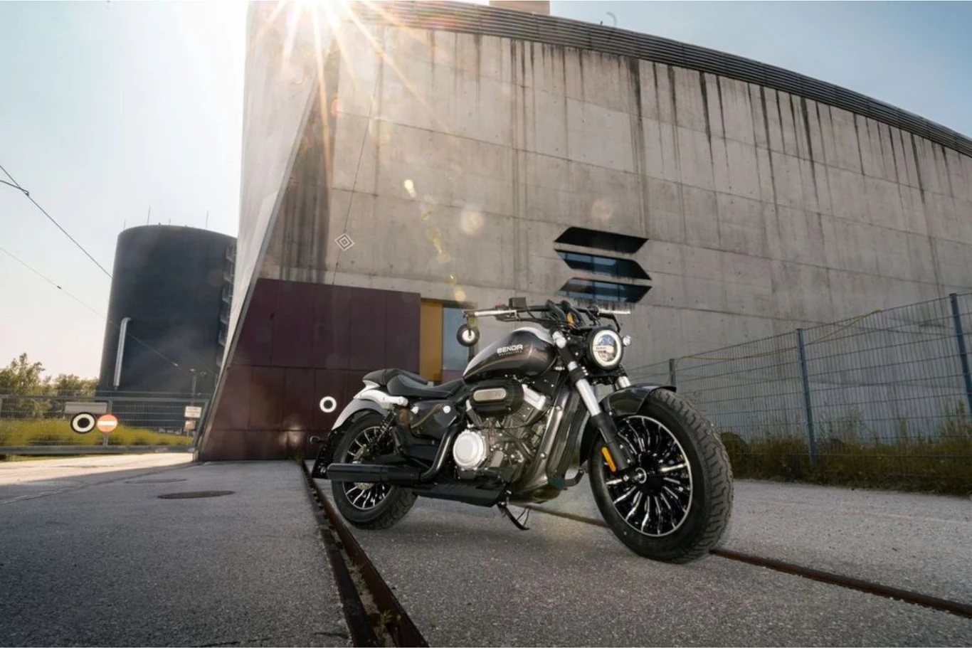 Czarny motocykl typu cruiser stoi na betonie przed nowoczesnym, betonowym budynkiem o surowej architekturze, w tle widoczne są metalowe ogrodzenie i promienie słońca wpadające z góry kadru.