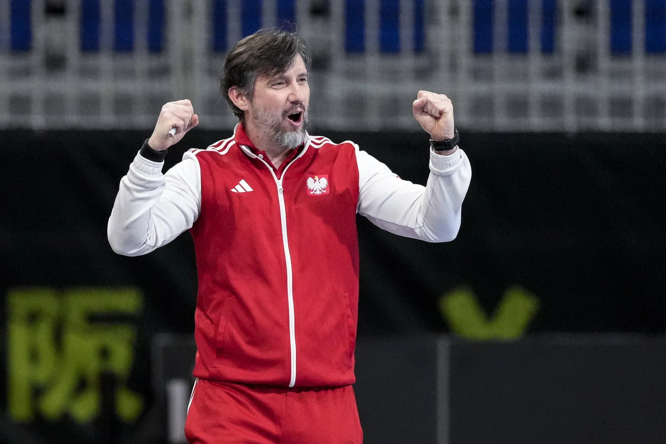 Mężczyzna w czerwonym dresie z biało-czerwoną flagą Polski na piersi unosi obie ręce w geście triumfu, z wyrazem entuzjazmu na twarzy.