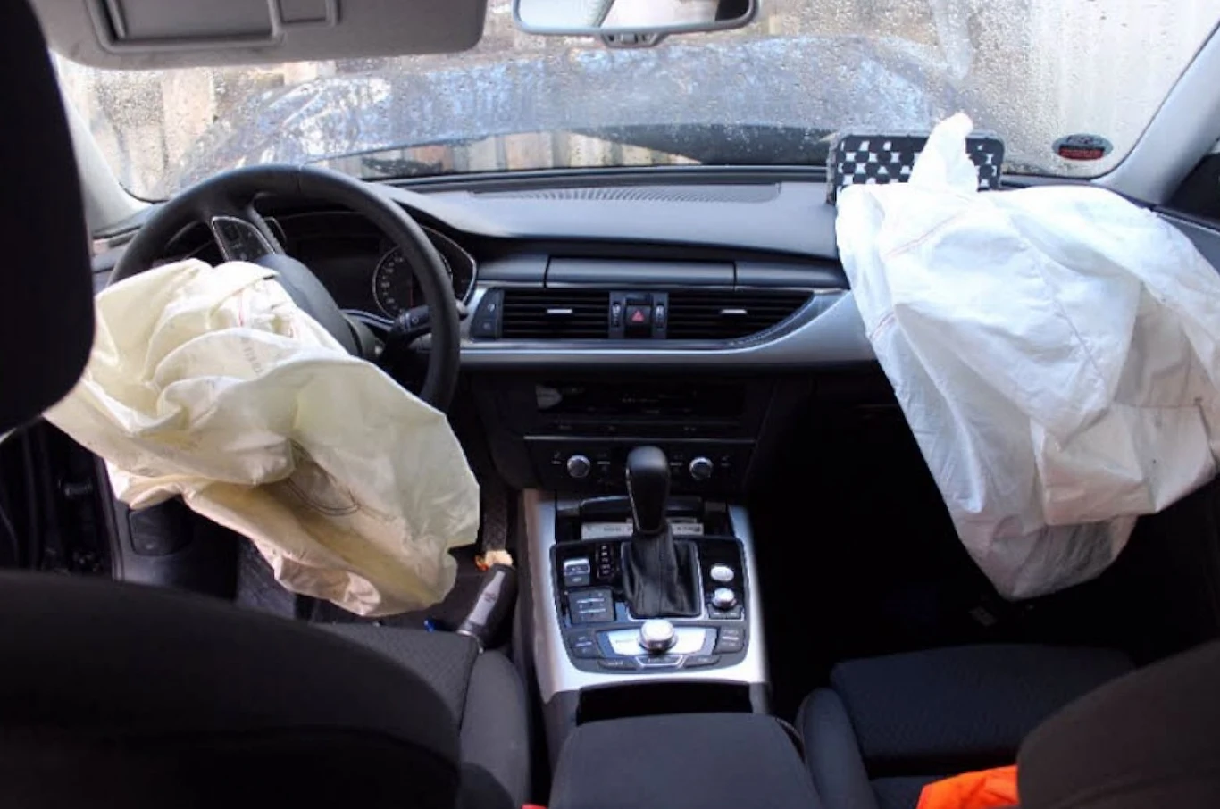 Wnętrze samochodu z wystrzelonymi poduszkami powietrznymi po stronie kierowcy i pasażera, widoczna konsola środkowa z dźwignią zmiany biegów oraz lekko zamglona szyba przednia.