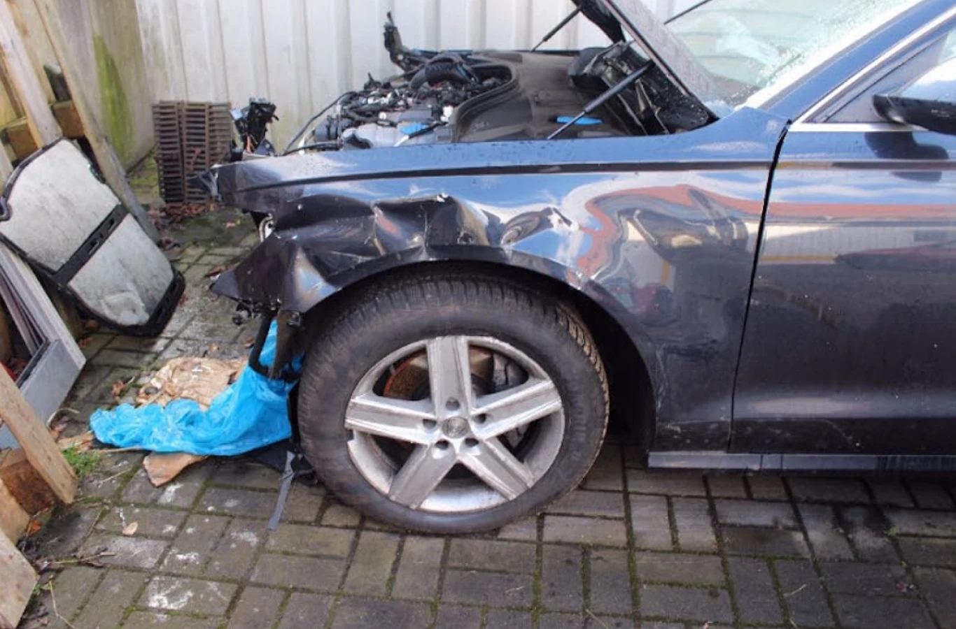 Mocno uszkodzony przedni lewy bok ciemnego samochodu z wgniecionym błotnikiem, widocznym uszkodzeniem maski i felgi, częściowo odsłonięty silnik.