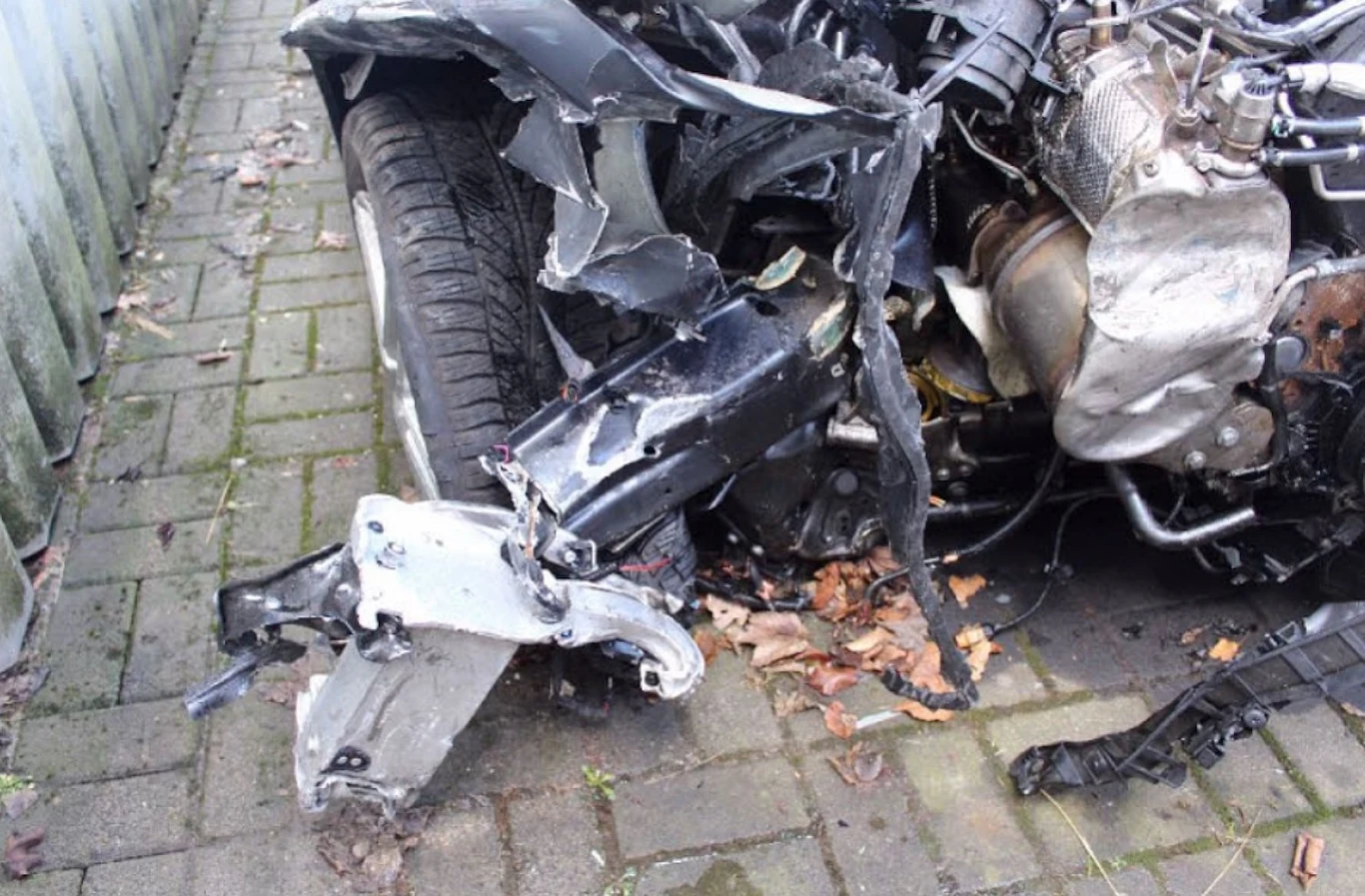 Zniszczony przód samochodu z widocznym uszkodzonym zawieszeniem i odsłoniętym silnikiem, leżące wokół fragmenty karoserii oraz liście na kostce brukowej.