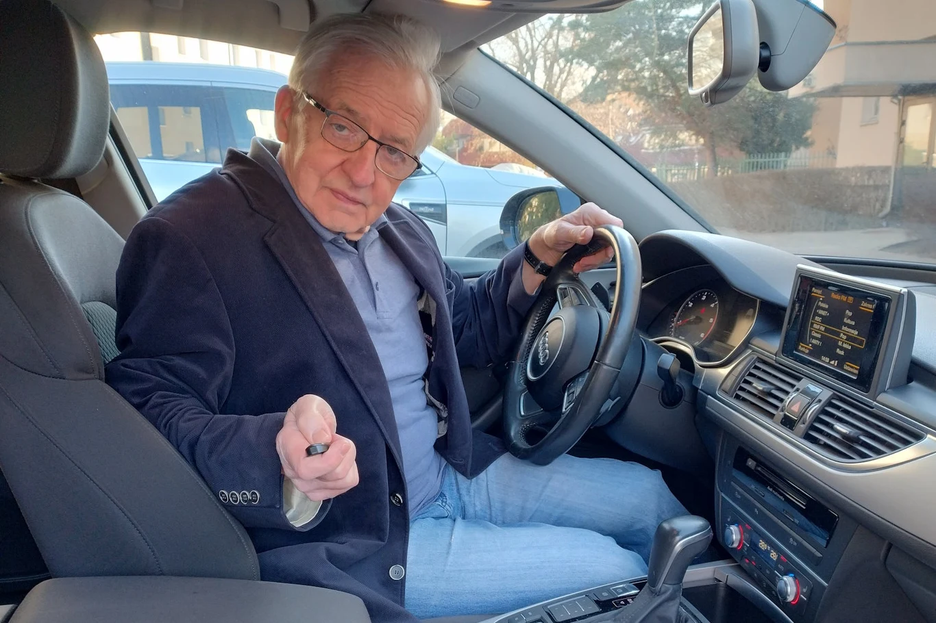 Starszy mężczyzna w okularach siedzi na miejscu kierowcy w samochodzie, trzymając w jednej ręce kluczyk, a drugą opiera na kierownicy; wnętrze auta nowoczesne, z widocznym wyświetlaczem i detalami deski rozdzielczej.