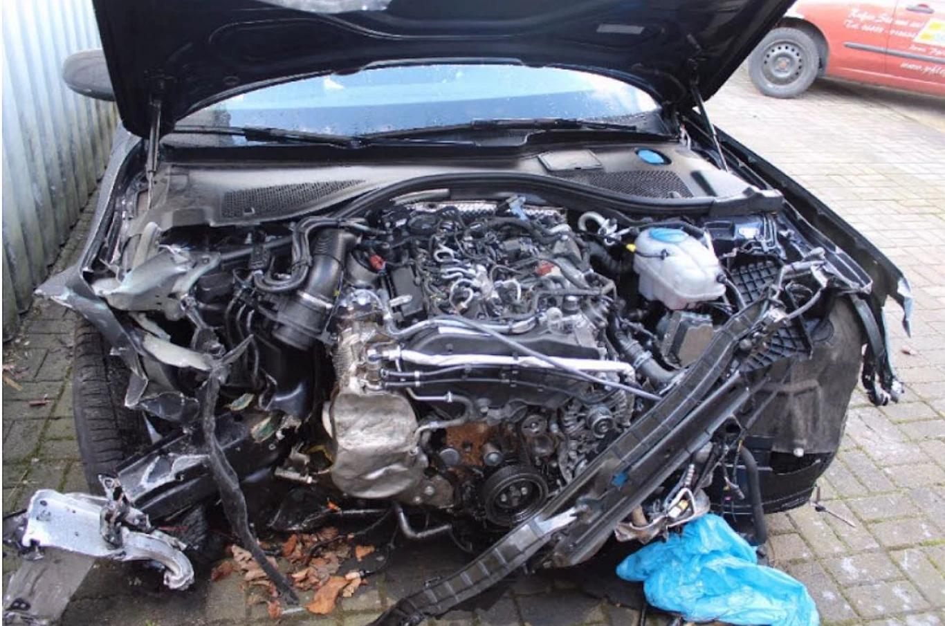 Samochód osobowy z poważnie uszkodzonym przodem, z widocznym silnikiem oraz elementami karoserii wygiętymi i odkształconymi wskutek silnego uderzenia, maska pojazdu otwarta, wokół wraku fragmenty zniszczonych części.